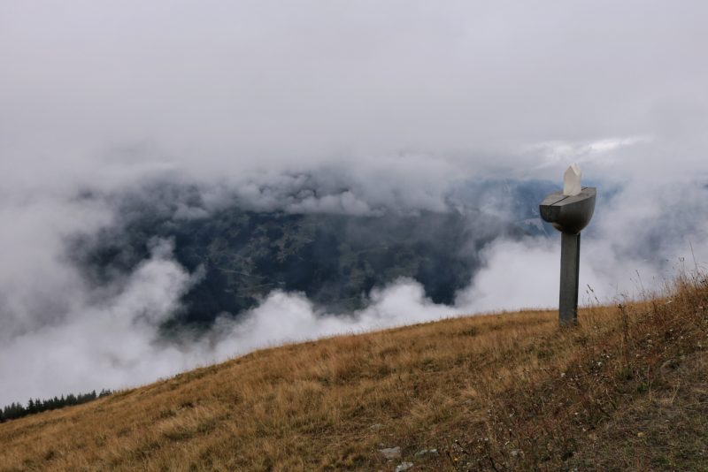 wooden pole with a boat shaped sculpture on top carrying a white model of a house. the scultpure is at the top of a grassy hill with thick clouds below and a green valley visible between the clouds.