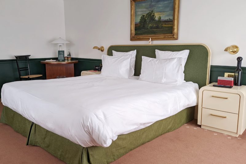 hotel room with pale red carpet  and cfream walls with a green bedframe with white sheets and a painting in a gold frame above the bed