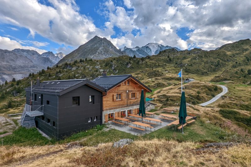 wooden two story bulding in traditional alpine style with a modern black cube shaped extension next to a dirt path surrounded by a grassy meadow with mountain peaks behind. verbier in summer.