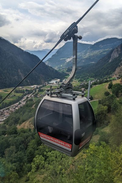 cable car with a red sign reading verbier 4vallees descending over a lush green valley in summer with mountains in the background