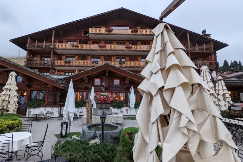exterior of a traditional swiss alpine chalet made from wood with a pointed roof and many balconies. there are tables with folder cream umbrellas on the terrace outside. verbier in summer.
