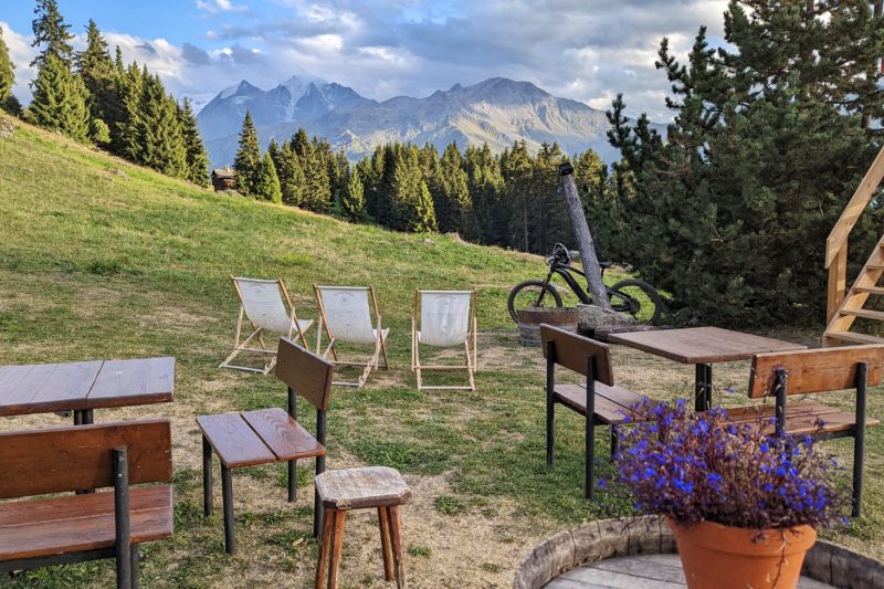outside terrace of Chez Dany in verbier on a summer evening with several wooden tables and stools and three deckchairs lined up facing a view of pine trees and distant moutains