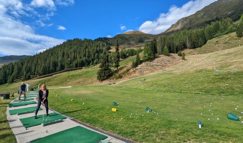 girl wearing black jeans and a black top swinging a golf club on a driving range with mountains and a pine forest in the background
