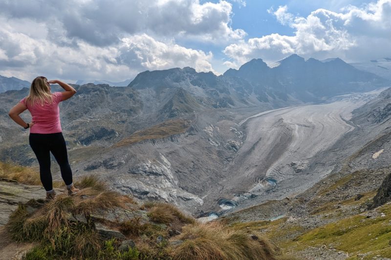blonde haired girl in a pink top and black leggings standing on the edge of a cliff looking out towards a grey coloured glacier with mountains in the background on a sunny summer day in verbier
