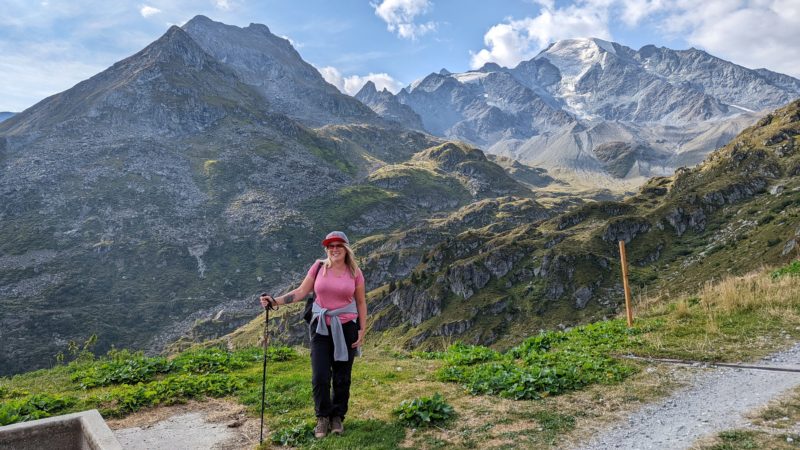 blonde haired girl wearing a grey and red cap, a pink t shirt, and black hiking trousers leaning on a hiking pole and standing on a patch of grass with tall grey mountains in the background - summer in verbier