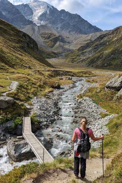 blonde haired girl wearing a pink t shirt, and black hiking trousers leaning on a hiking pole standing next to a river with a small wooden footbridge crossing it and a tall grey mountain in front of her with snow on the peak