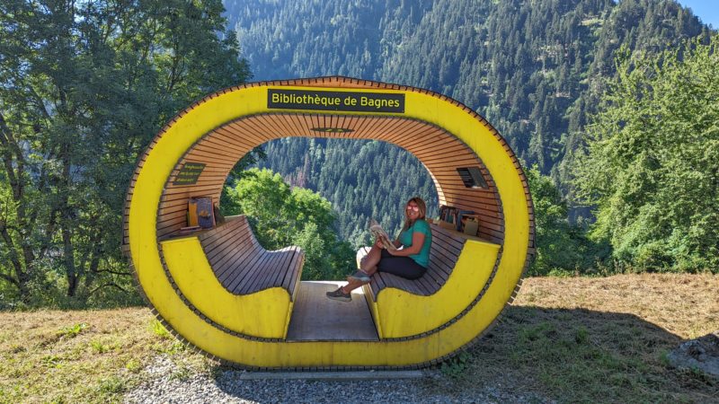 blonde girl wearing black shorts and a bright green t shirt sitting in an oval shaped wooden yellow hut with empty sides so that the pine forest behind is visible through the hut.