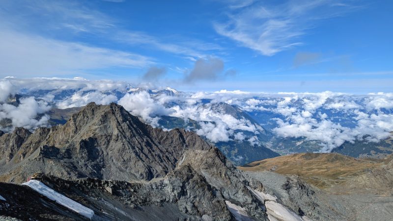 landscape view of a rocky mountainside with several snowy peaks and fluffy clouds behind it on a sunny summer day with a bright blue sky in verbier