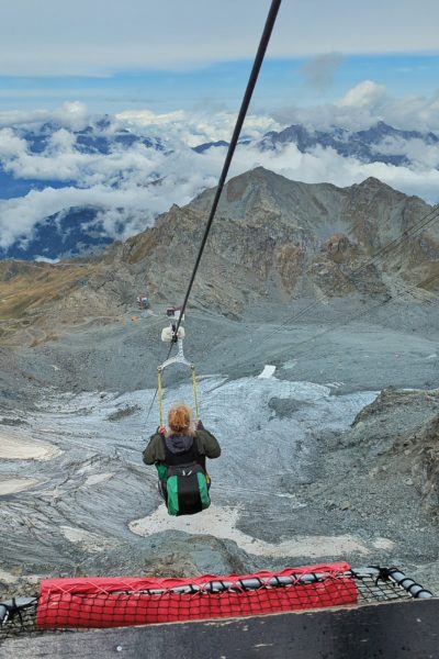 lady with strawberry blonde hair wearing a grey jacket and handing from a zipline in a sitting position travelling downhill above a grey glacier and rocky landscape with mountains behind. verbier in summer.