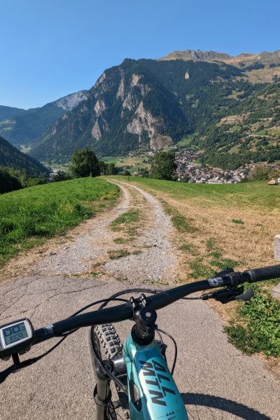 handlebars of a turquoise mountain bike at the top of a gravel path leading downhill between grass with a green forest covered mountain peak in the distance