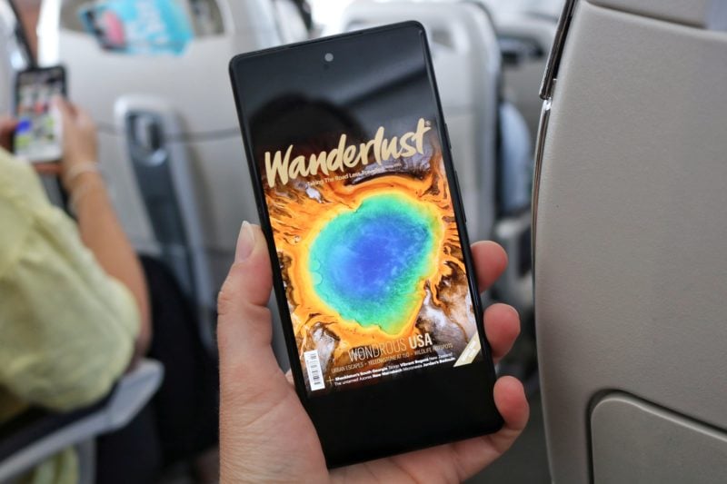 Hand holding a mobile phone in front of the aisle of an aeroplane with the cover of Wanderlust magazine visible on the phone screen. Readly magazine reader app review. 