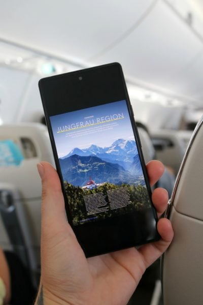hand holding a phone with a magazine article about the Jungfrau Region open on the screen. the phone is held up in front of the aisle of an aeroplane with grey seatbacks visible behind. 