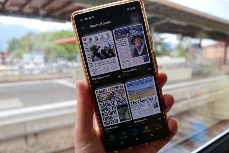 hand holding a phone with four newspaper covers displayed in a grid. the phone is in front of a train window with tracks and a station out of focus behind it.