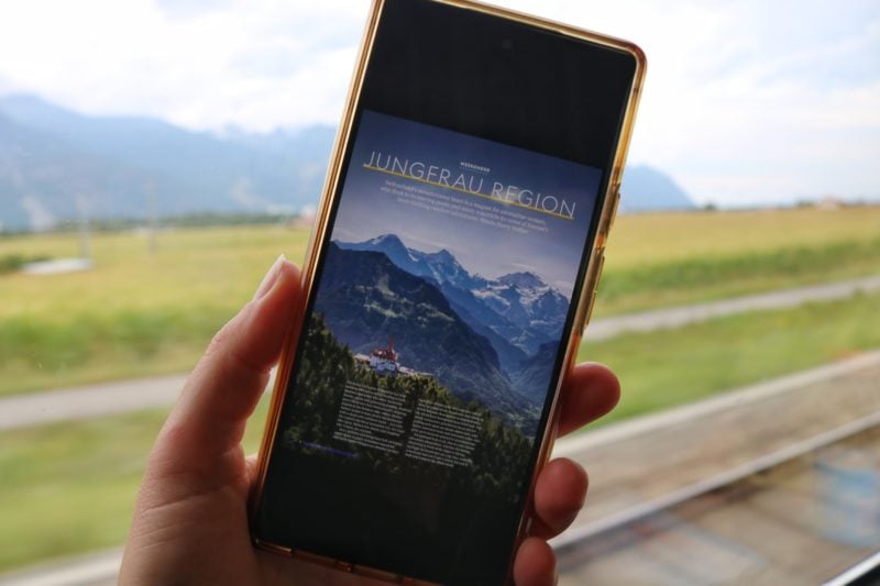 hand holding a phone with a magazine article about the Jungfrau Region open on the screen. the phone is being held in front of a dirty train window with train tracks and grass behind and some out of focus mountains in the distance. 