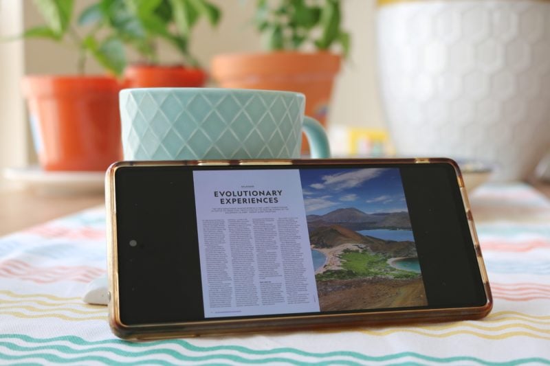 close up of a phone with a double page magazine open on the screen. the phone is propped against a turquoise coffee mug with some pot plants in the background. 