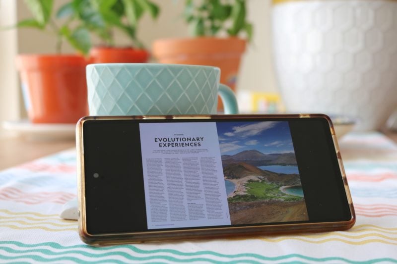 close up of a phone with a double page magazine open on the screen. the phone is propped against a turquoise coffee mug with some pot plants in the background.