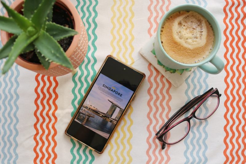 flat lay of a table cloth with multicoloured wiggly lines. from left to right on top of the tablecloth there is an aloe vera plant in an orange pot, a smartphone with an article about singapore open, a turquoise mug full of coffee, and a pair of glasses with purple frames