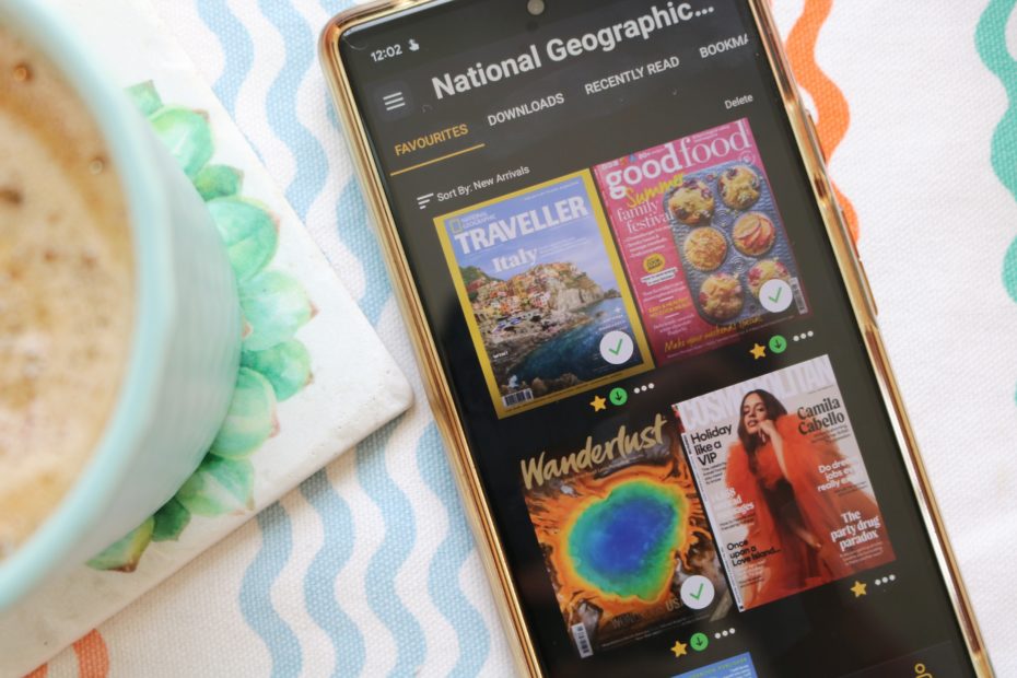 Flatlay of the Readly App open on a smartphone with four magazine covers in a grid. The phone is on a white tablecloth with multicoloured wiggly lines and the edge of a cup of coffee is visible on the left. Readly Review.