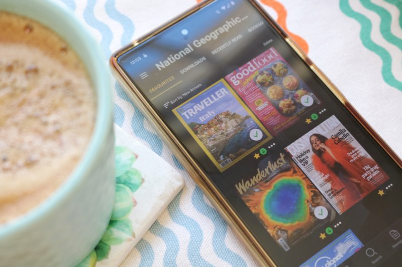 Flatlay of the Readly App open on a smartphone with four magazine covers in a grid. The phone is on a white tablecloth with multicoloured wiggly lines and the edge of a cup of coffee is visible on the left. Readly Review and promo code