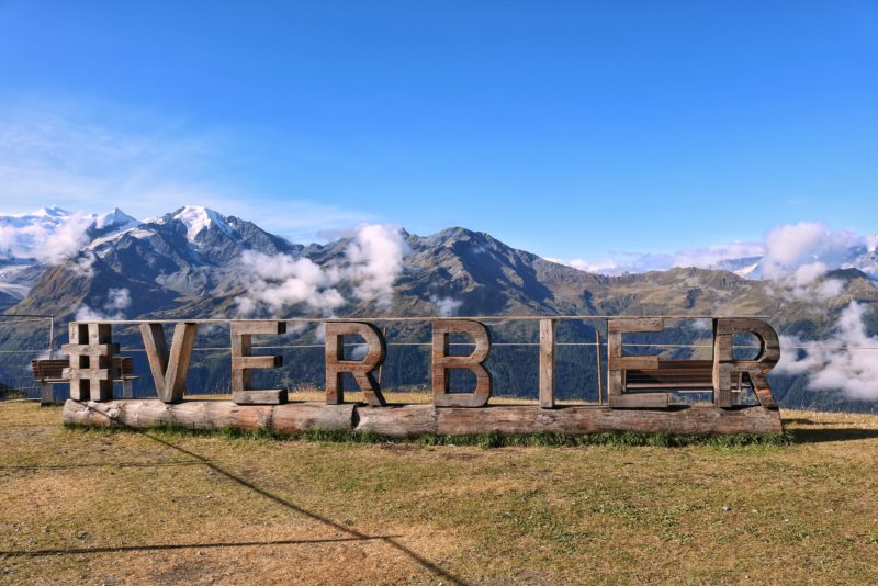 large wooden Verbier sign on a grassy lawn with snow capped mountains in the background and an empty blue sky overhead in summer