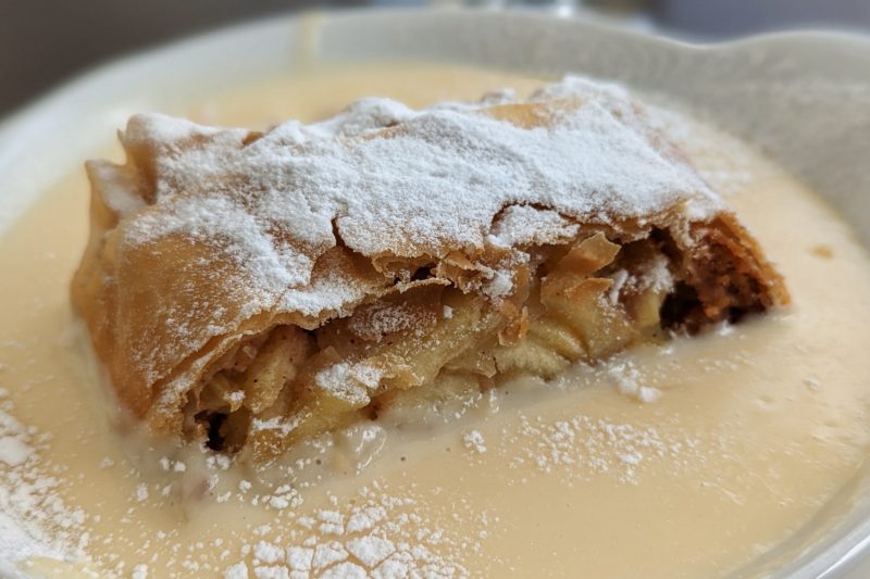 close up of a roll of pastry with chopped apple inside itopped with white powdered sugar and surrounded by pale yellow custard