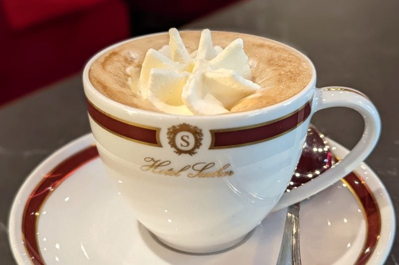 close up of a small china cup and saucer printed wiht Hotel Sacher in gold letters and filled with coffee topped with a squirt of whipped cream