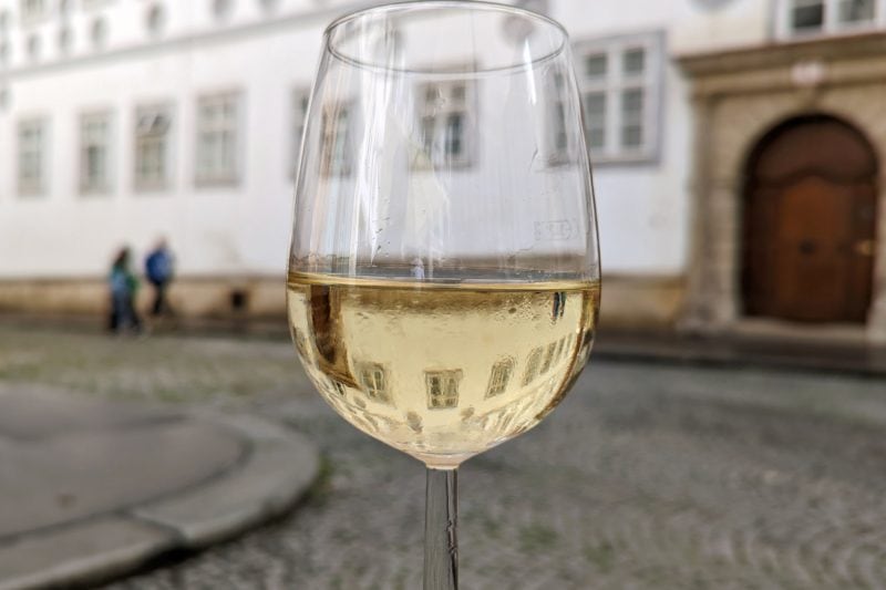 close up of a wine glass filled with slightly yellow white wine in front of a cobblestone square in vienna with a white building in the background with a large dark brown wooden door in a stone frame. 