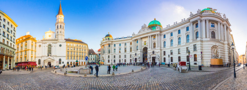 panorama of a large white palace on a cobbled square in vienna beside a white church tower on a very sunny day with blue sky above. What to eat in Vienna. 