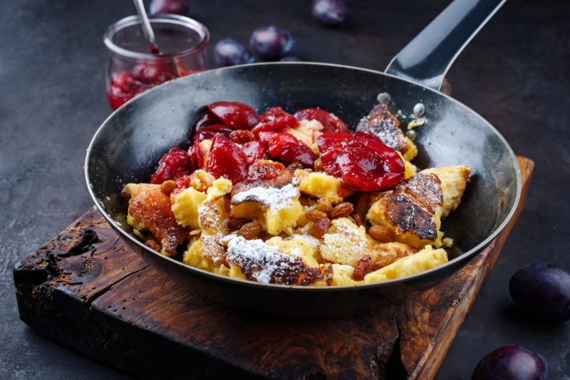close up of traditional Austrian Kaiserschmarren shredded pieces of yellow pancake topped with powdered sugar and plums in a rustic black frying pan on a wooden sideboard. what to eat in vienna. 
