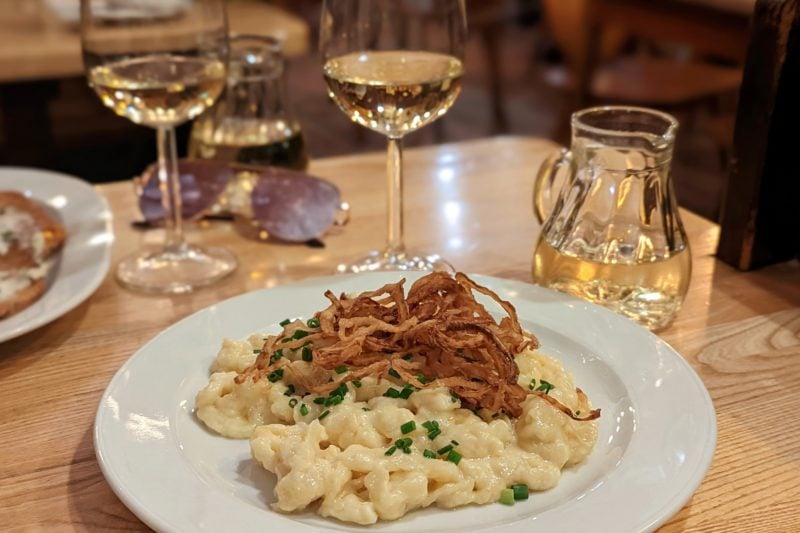 white plate on a light wooden table with a large serving of pasta in a cheese sauce topped with crunchy fried brown onions. there are two wine glasses on the table filled with white wine and a small jug of white wine beside them.