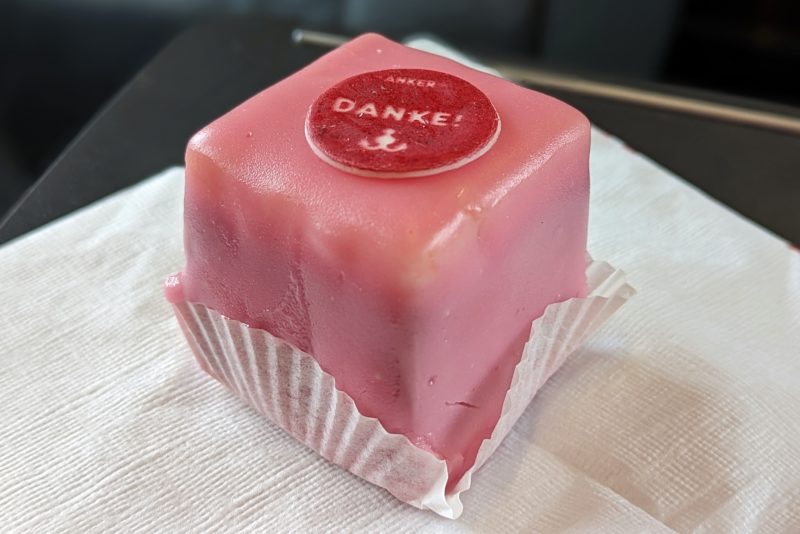close up of a small cube shaped cake covered in pink ice cream with a red sticker on top that says Danke. the cake is on a white napkin. Punschkrapfen is a traditional dessert in viennese cuisine