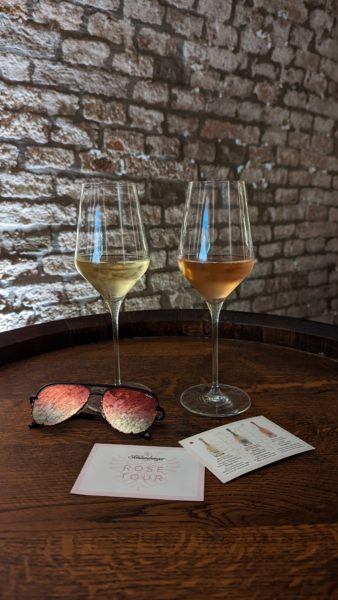 wo wine glasses filled with one white and one rose champagne next to a pair of pink sunglasses on top of a wooden barrel and in front of a stone cellar wall