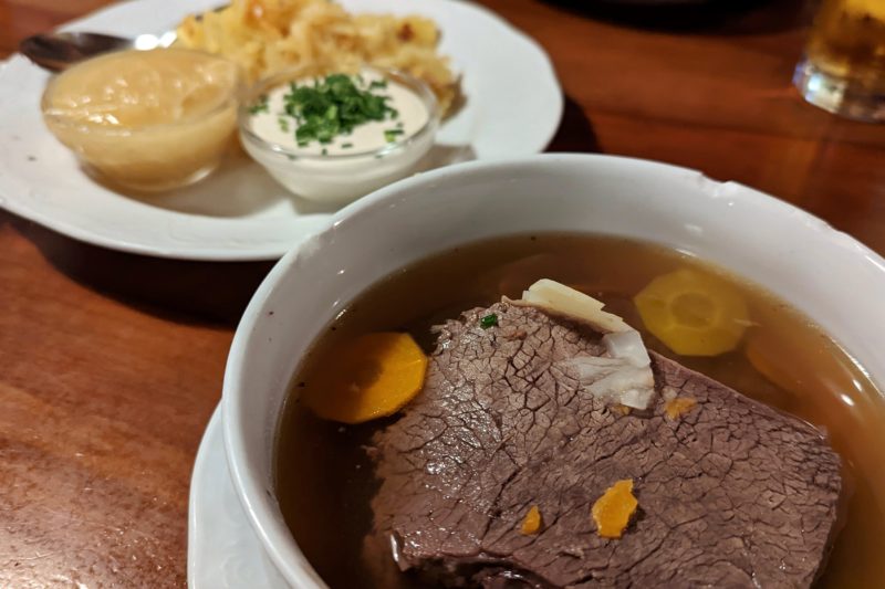 white bowl on a brown wooden table containing a watery broth and large pieces of beef with a few carrot circles. on another white plate behind is some potatoe and two small bowls with one white and one beige coloured sauce. tafelspitz is a traditional viennese food. 