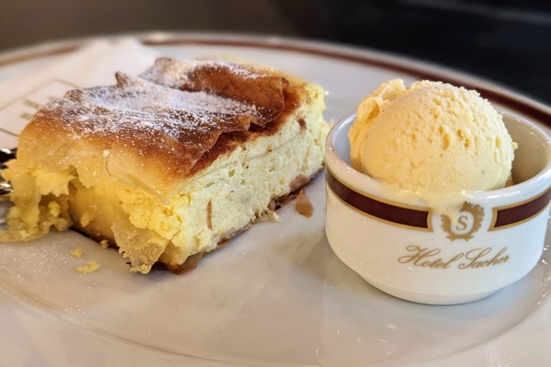 close up of a white plate with a piece of dense creamy yellow cake topped with flaky pastry. there is a small white bowl printed with Hotel Sacher in gold letters containing a scoop of yellow ice cream. at cafe sacher vienna.