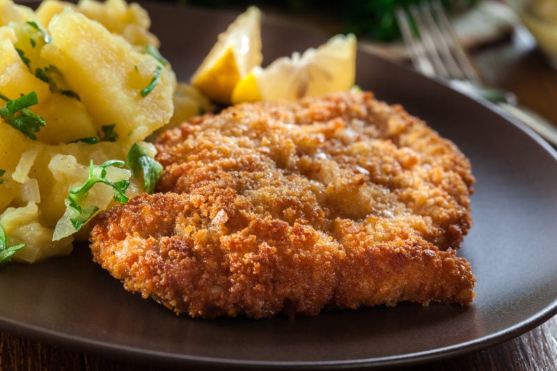 close up of a breaded viennese schnitzel joint on a black plate with lemon wedges and potatoes just out of focus behind.
