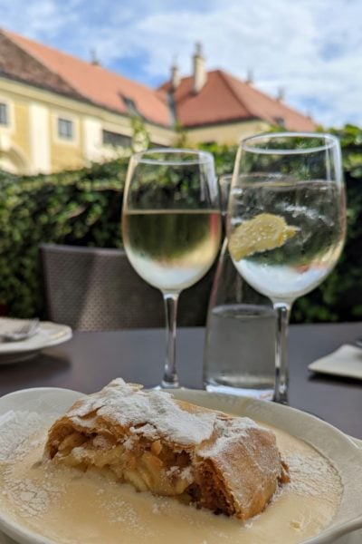 two wine glasses with spritzers on a grey table with a bowl in front containing apple strudel and pale yellow custard. behind is a green hedge and a yellow building with red tiled roof on a very sunny day