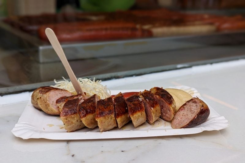 white paper plate on a white marble countertop with a brown cooked sausage cut into small slices and a wooden fork sticking up from the sausage at a wurstelstand in vienna