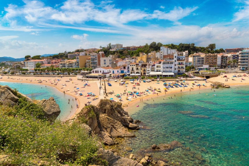 Standing on a hilltop on a headland looking back towards the mainland at a long white sandy beach covered in tourists with a large town of mainly white buildings behind. Blanes in Costa Brava Spain