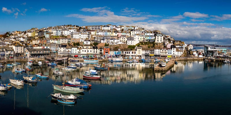 looking across a very calm blue harbour with many fishing boats in it at a town on a hillside with many colourful buildings reflected in the still water. brixham in devon england
