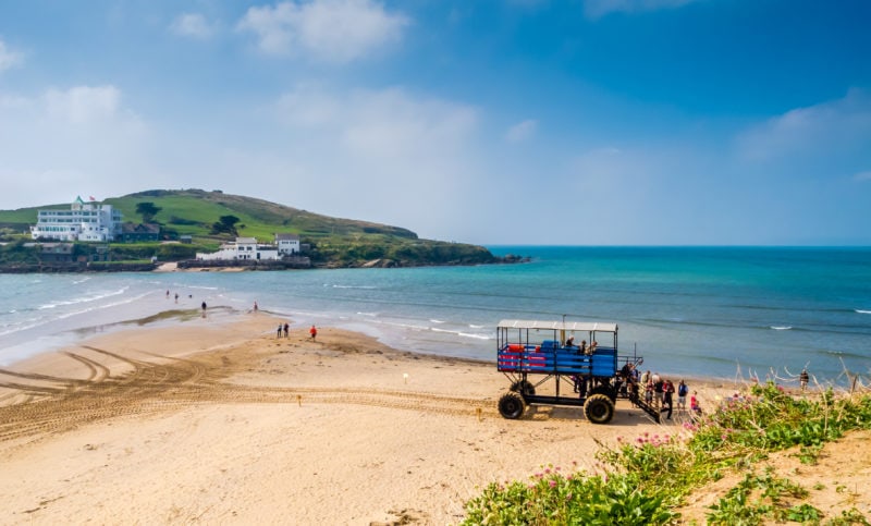 white sandy beach with a blue tractor on it and a thin strip of sand leading to a small tidal island with a white building on it. burgh island is one of the best places to visit in south devon.