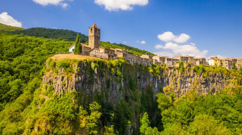 town with lots of houses built from beige stone on a clifftop surrounded by greenery with low green mountains behind