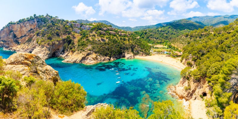 View from a low clifftop looking down at a bright turquoise bay with a small sandy beach and a low hill covered in green foliage on the far side - Cala Giverola, one of the best places to visit in Costa Brava Spain