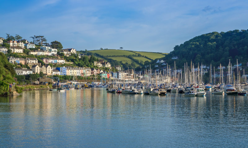 blue river full of yachts with green hills in the background and a town on either side of the river. there are colourful houses at kingswear on the left while the town on the right is in shadow. best places to visit in south devon.