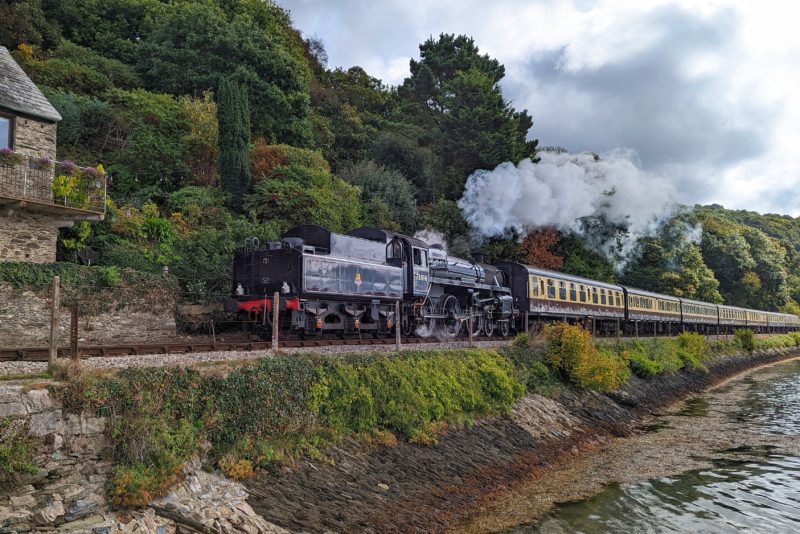 black steam train engine going along a track beside a river with thick green woodland behind it