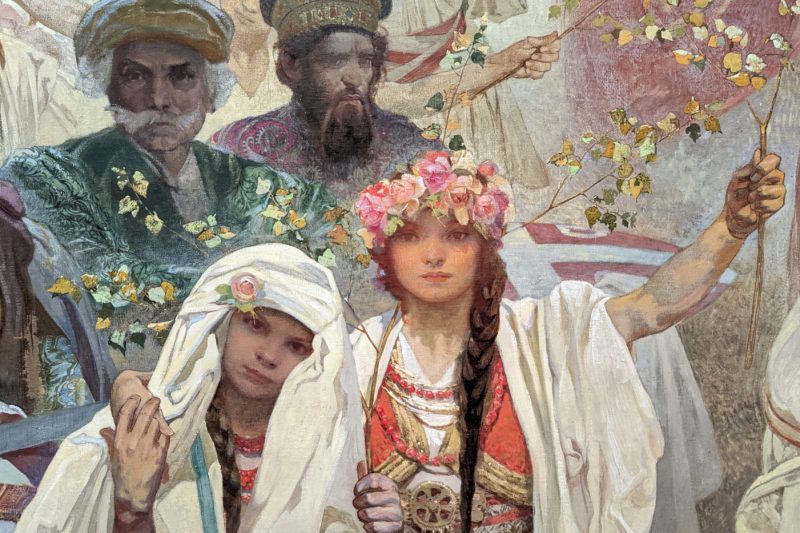 close up of a portion of a painting showing two young girls in traditional czech costume with white gowns and brown hair in long plaits. The taller girl haas pink flowers in her hair. Day trips from Brno. 