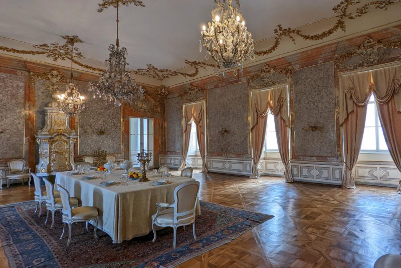 Interior or a room with parquet floor and marble walls. there is a long table with a white tablecloth and expensive looking china dishes. three chandeliers are hanging from the white cieling. inside castle valtice. 