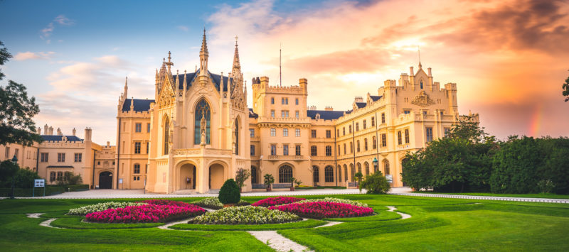 Lednice Chateau with beautiful gardens and parks on sunny summer day. There is a flower bed with red flowers in the foreground and an orangey sunset with a rainbow in the background. Best day trips from Brno South Moravia. 