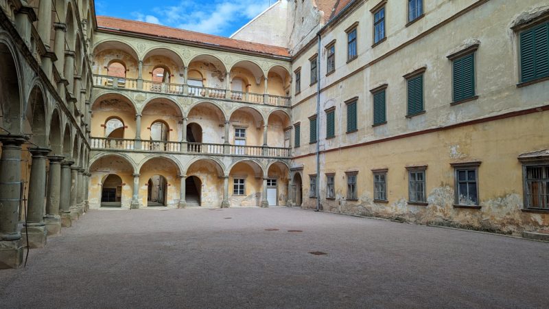 Courtyard surrounded by yellow stone walls with many windows. The back wall has 3 tiers of collonades and archways. Moravský Krumlov Castle.