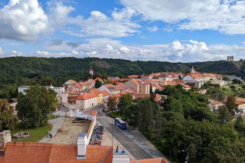 Looking out from the top of a tower at a small town where all the houses have orange tiled roofs surrounded by thick green forest with a green hill in the background on a sunny day with blue sky. Moravský Krumlov near Brno in South Moravia. 