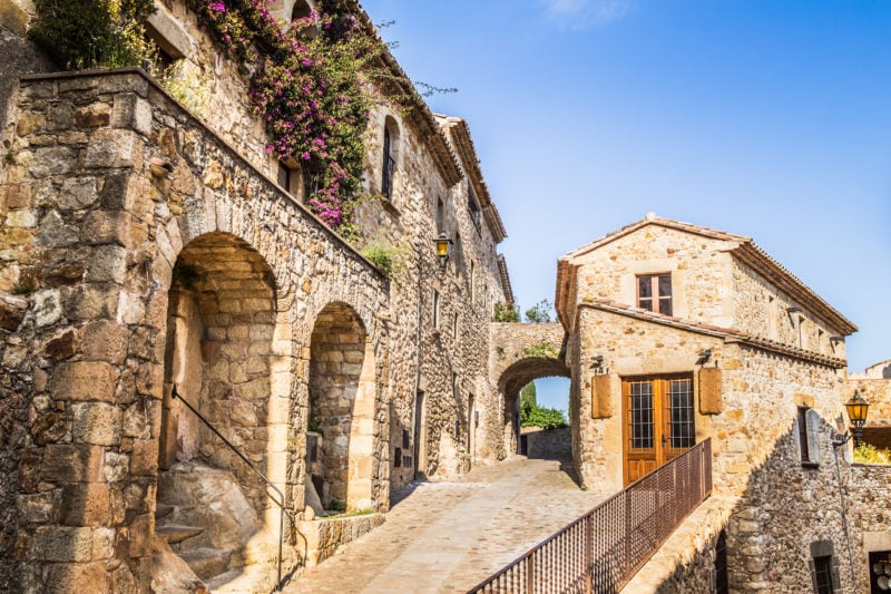 medieval buildings in a village centre built from beige coloured stone with a cobbled street running past some stone archways on a sunny day with a clear blue sky - pals is one of the best places to visit in costa brava